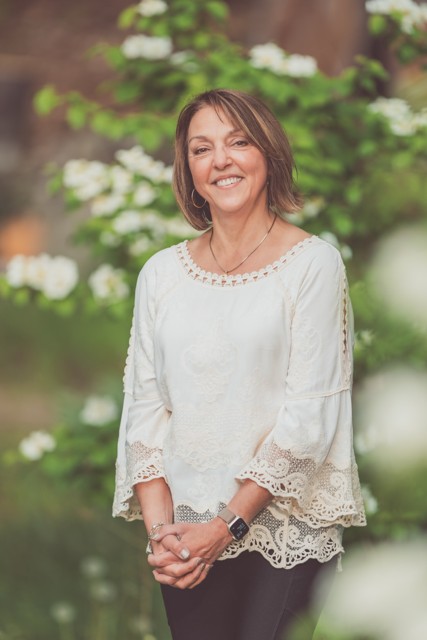 Judy Morin, founder and curator of Gather Wellness Center, standing outdoors in front of flowering shrubs in a white embroidered blouse.